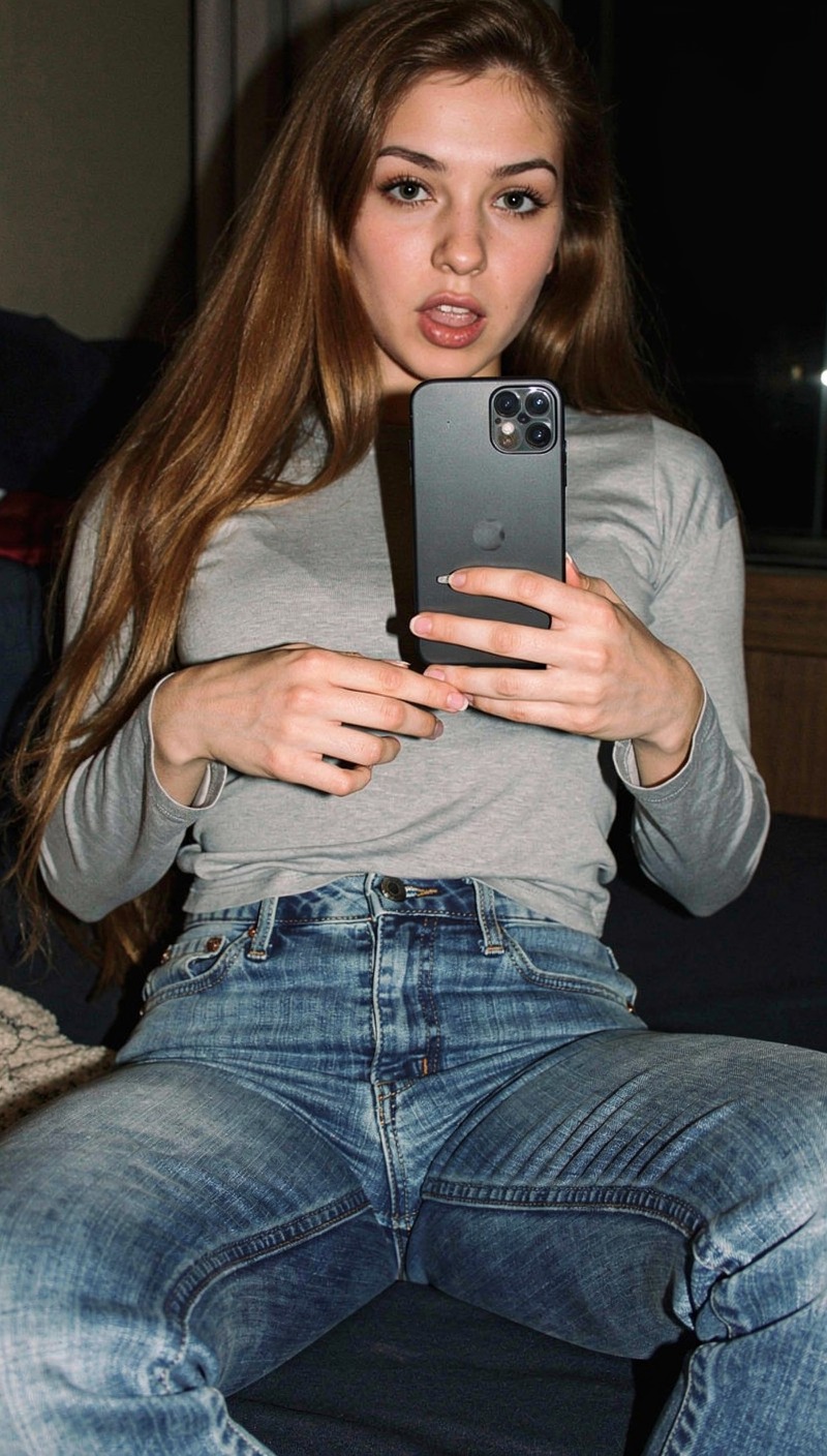 A young woman sits casually on a couch, holding a smartphone in both hands while taking a mirror-style photo and looking directly at the camera. The image is captured in a candid, modern lifestyle photography style with natural skin tones and sharp focus. She appears indoors in a dimly lit, intimate setting, wearing casual clothing, with a relaxed, informal mood that suggests a spontaneous, personal moment.